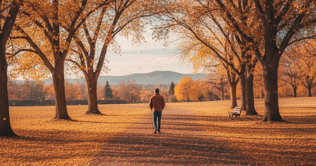 A person seen from behind walking alone on a tree-lined park path in autumn with foothills in the distance